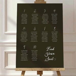 May include: A dark brown seating chart on an easel with white calligraphy. The chart is divided into ten sections, each with a number and a list of names. The words "Find Your Seat" are written in elegant script.