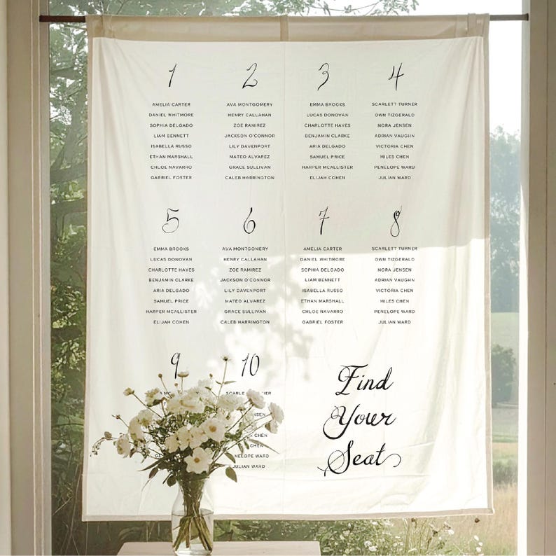 May include: A cream-colored fabric wedding seating chart with elegant calligraphy. The chart is divided into numbered sections with guest names. The phrase "Find Your Seat" is written in cursive at the bottom. A vase of white flowers sits below.