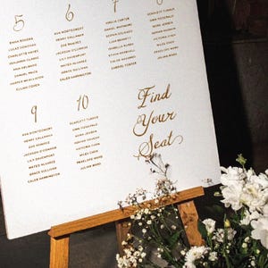 May include: A white seating chart with gold lettering and guest names, displayed on a wooden easel. The chart is decorated with white flowers and greenery. The text "Find Your Seat" is prominently displayed.