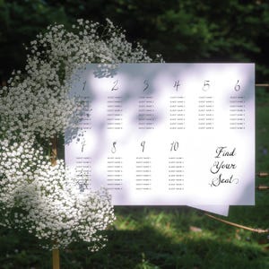 May include: A white seating chart with handwritten numbers and guest names. The text "Find Your Seat" is written in elegant script. The chart is displayed outdoors with white flowers.