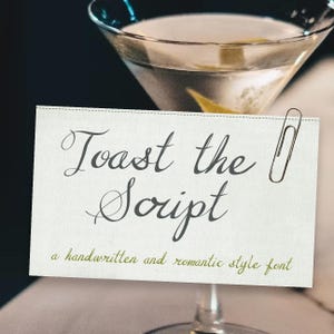May include: A martini glass with a clear liquid and a lemon twist. A white card with the words "Toast the Script" in elegant script font is attached with a paper clip. The card also reads "a handwritten and romantic style font."