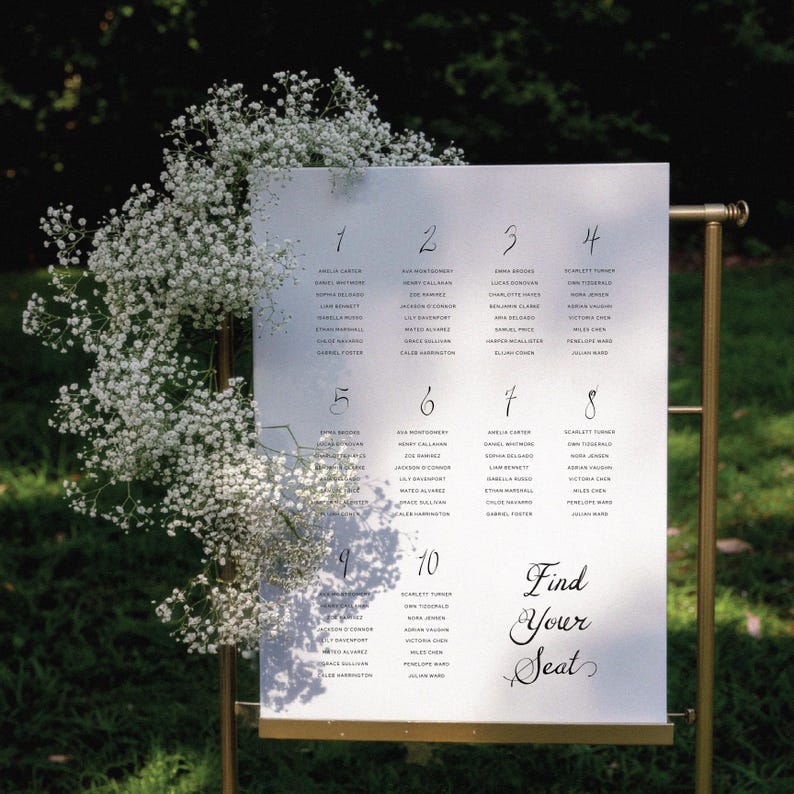 May include: A wedding seating chart with a gold metal frame. The white sign displays guest names organized by table number. Delicate white flowers cascade down the side. The text "Find Your Seat" is written in elegant script.