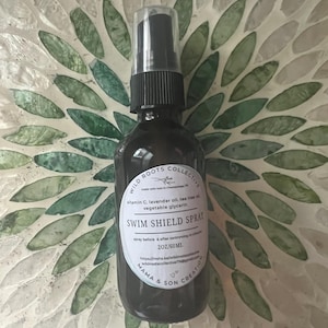 Puede incluir: Un frasco rociador de vidrio marrón oscuro con una tapa negra y una etiqueta blanca. La etiqueta dice "SWIM SHIELD SPRAY" y enumera los ingredientes. La botella es de 60 ml. El fondo es un patrón de mosaico con baldosas verdes y blancas en forma de hoja.