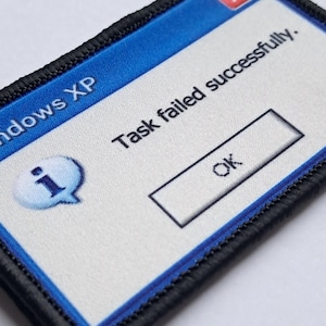 Könnte beinhalten: Ein rechteckiger Aufnäher mit schwarzem Rand und blau-weißem Design. Der Aufnäher zeigt eine Fehlermeldung im Windows XP-Stil mit der Aufschrift "Task failed successfully." mit einer "OK"-Schaltfläche.