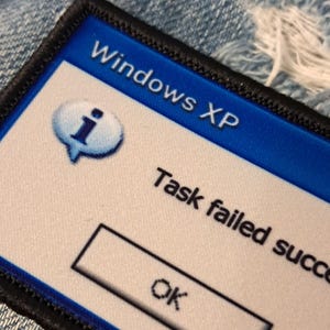 Pu&ograve; includere: Una toppa rettangolare con bordo nero. Presenta uno sfondo blu con la scritta "Windows XP". La toppa include una nuvoletta con una "i" all'interno e la scritta "Task failed success" con un pulsante "OK".