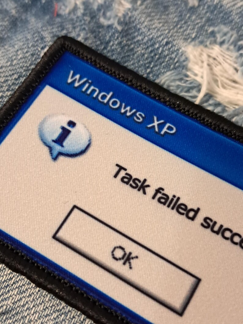 Pu&ograve; includere: Una toppa rettangolare con bordo nero. Presenta uno sfondo blu con la scritta "Windows XP". La toppa include una nuvoletta con una "i" all'interno e la scritta "Task failed success" con un pulsante "OK".