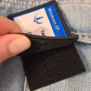 Pu&ograve; includere: Una toppa nera e blu con la scritta "Windows XP" e un'icona di informazione. La toppa &egrave; attaccata a un tessuto denim con un retro in velcro nero.