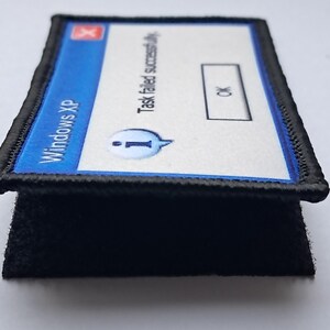 Pu&ograve; includere: Una toppa rettangolare con un messaggio di errore di Windows XP. Il design blu e bianco recita "Task failed successfully" con un pulsante "OK". La toppa ha un bordo nero e un retro in velcro nero.