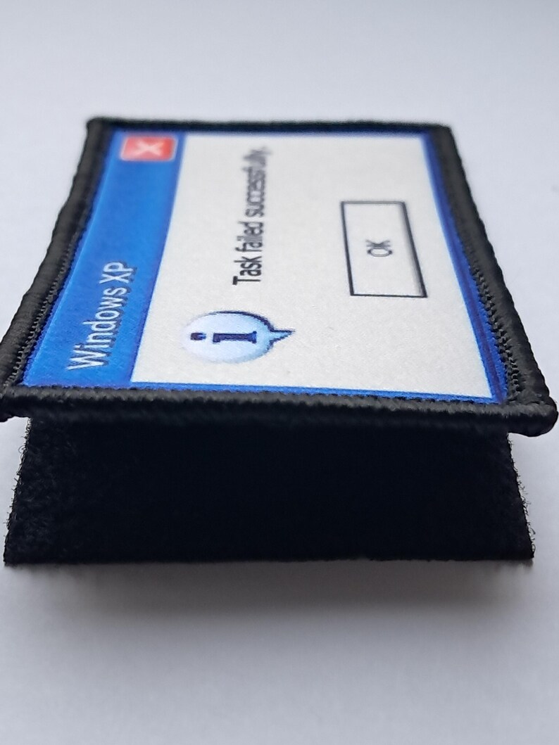 Pu&ograve; includere: Una toppa rettangolare con un messaggio di errore di Windows XP. Il design blu e bianco recita "Task failed successfully" con un pulsante "OK". La toppa ha un bordo nero e un retro in velcro nero.