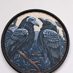 Könnte beinhalten: Runder Aufnäher mit zwei schwarzen Raben mit blauen und weißen Details, vor einem blau-braunen Hintergrund. Der Aufnäher hat einen schwarzen Rand und ist für Kleidung oder Accessoires geeignet.