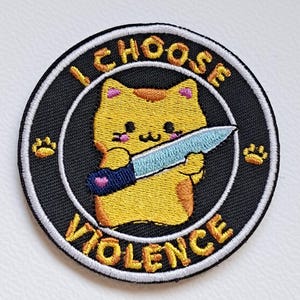 Può includere: Toppa ricamata con un gatto dei cartoni animati che tiene un coltello. Il gatto è giallo con guance rosa e un cuore sul manico del coltello. La toppa è rotonda con la scritta "I CHOOSE VIOLENCE" in lettere gialle su sfondo nero.