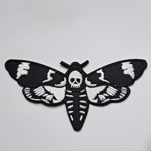 May include: Black and white embroidered patch in the shape of a moth. The moth features a skull design on its head and a skeleton-like pattern on its body. The wings have white accents.