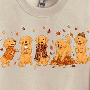 Könnte beinhalten: Ein beigefarbenes Sweatshirt mit Rundhalsausschnitt und einem Grafikdruck von fünf Golden Retrievern. Die Hunde sind in verschiedenen herbstlichen Posen dargestellt, tragen einen Pullover, einen Schal und einen Eichelhut, umgeben von Blättern und Sternen.