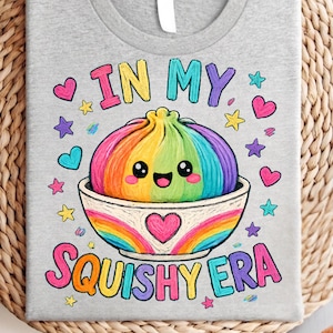 Könnte beinhalten: Ein graues T-Shirt mit einer bunten Grafik eines regenbogenfarbenen Knödels in einer Schüssel mit einem lächelnden Gesicht. Der Text lautet IN MY SQUISHY ERA, umgeben von kleinen rosa, blauen und gelben Herzen und Sternen in leuchtenden Farben.