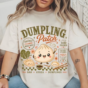 Könnte beinhalten: Ein cremefarbenes T-Shirt mit einem Grafikdruck mit dem Text DUMPLING Patch, Freshly Steamed Made Daily, Hand Folded und Bao Gyoza Dumplings. Das Design zeigt eine Cartoon-Teigtaschenfigur, kleinere Teigtaschen, eine Schüssel und Blätter.