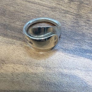 May include: A silver ring crafted from a spoon, featuring a curved design. The ring has a polished finish and a unique, handcrafted appearance. The ring is displayed on a wooden surface.