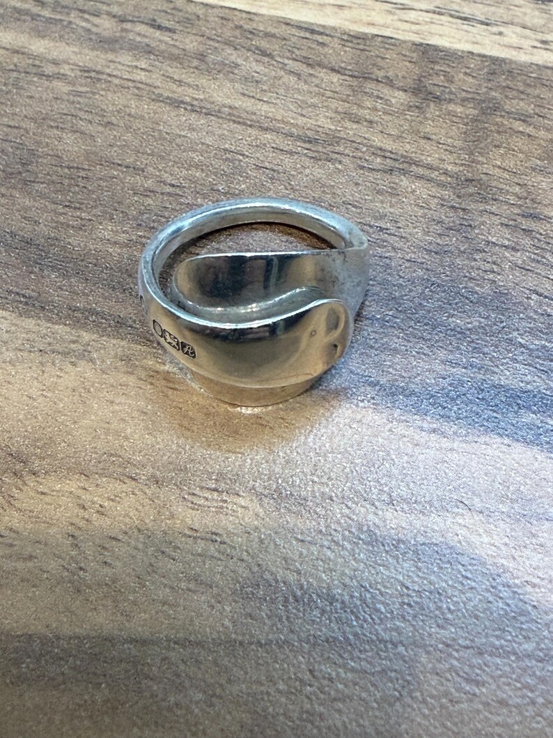 May include: A silver ring crafted from a spoon, featuring a curved design. The ring has a polished finish and a unique, handcrafted appearance. The ring is displayed on a wooden surface.
