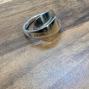 May include: A silver ring with a unique design, possibly handcrafted. The ring has a wide, curved band and a rounded, textured centerpiece. The metal has a polished finish, reflecting light. The ring appears to be a size 7.