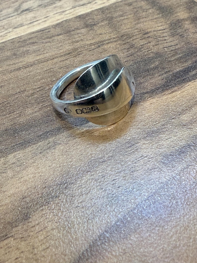 May include: A silver ring with a unique design, possibly handcrafted. The ring has a wide, curved band and a rounded, textured centerpiece. The metal has a polished finish, reflecting light. The ring appears to be a size 7.