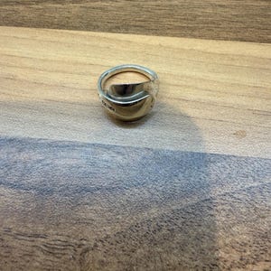 May include: A silver ring crafted from a spoon, featuring a curved bowl and a band. The ring has a polished finish and is set against a wooden surface. The ring's design is unique and handmade.