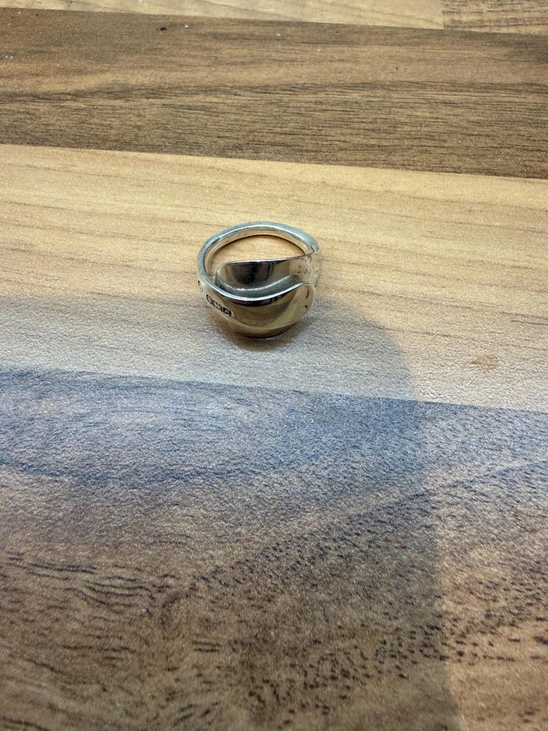 May include: A silver ring crafted from a spoon, featuring a curved bowl and a band. The ring has a polished finish and is set against a wooden surface. The ring's design is unique and handmade.