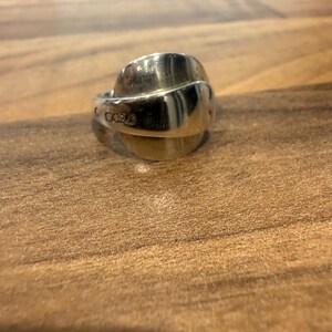 May include: A silver ring with a spoon-like design. The ring has a curved, rounded top and a band that wraps around the finger. The ring is made of a shiny, reflective metal and is set against a wooden surface.