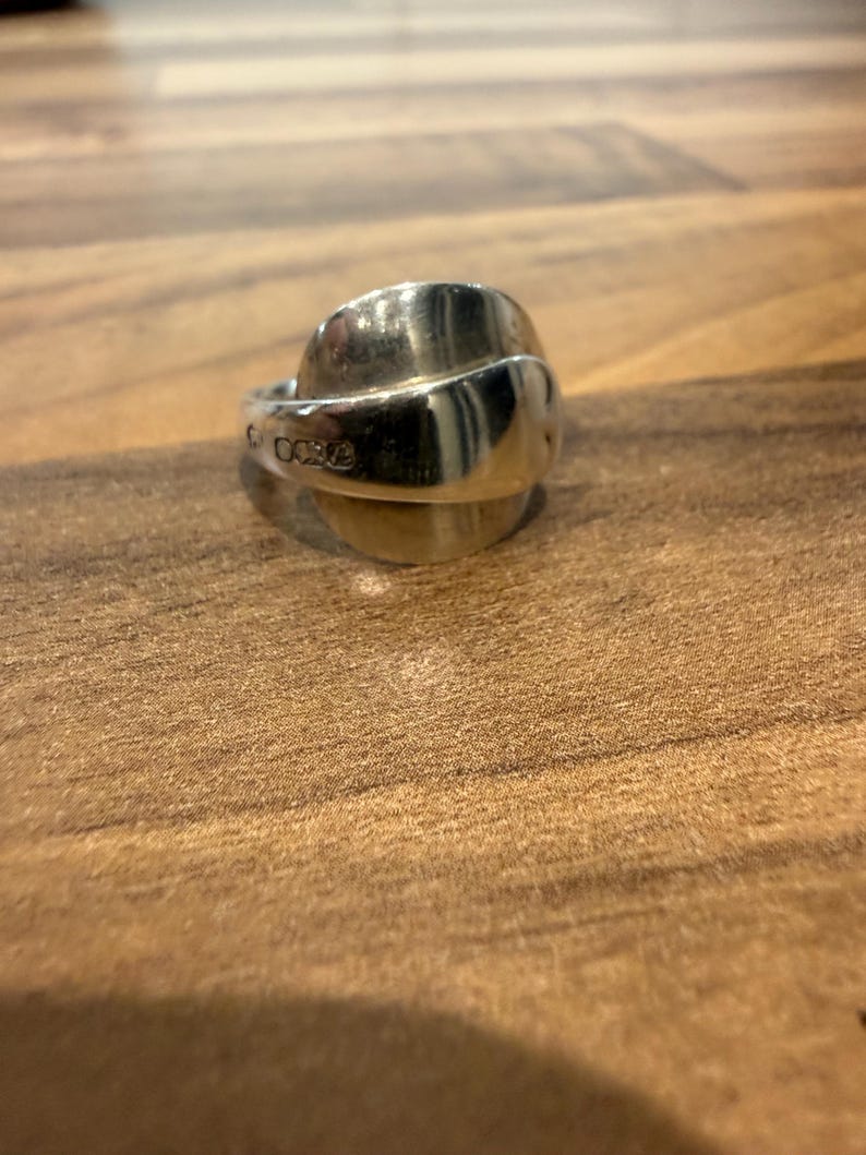 May include: A silver ring with a spoon-like design. The ring has a curved, rounded top and a band that wraps around the finger. The ring is made of a shiny, reflective metal and is set against a wooden surface.
