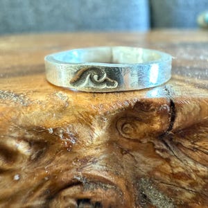 Może przedstawiać: Srebrny pierścionek z falistym wzorem na obrączce. Pierścionek leży na kawałku drewna.