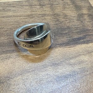 May include: A silver ring crafted from a spoon, featuring a curved design. The ring has a polished finish and visible markings on the band. The ring is a unique piece of jewelry.