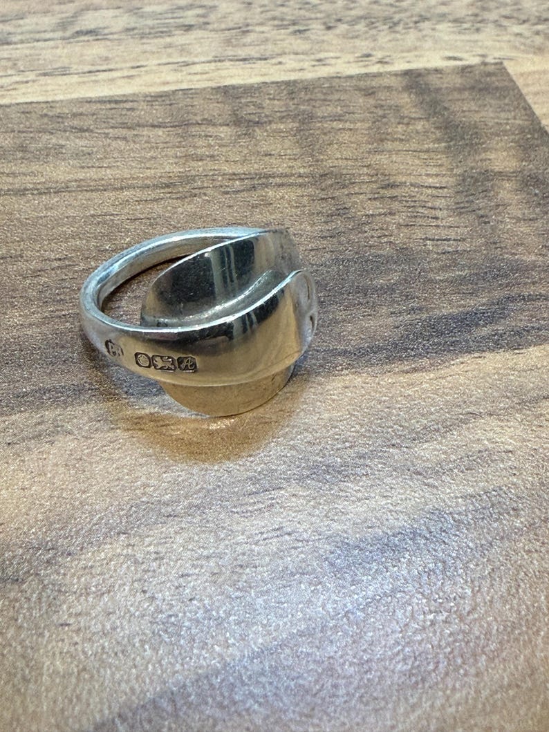 May include: A silver ring crafted from a spoon, featuring a curved design. The ring has a polished finish and visible markings on the band. The ring is a unique piece of jewelry.