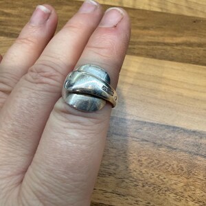 May include: A silver ring crafted from a spoon, featuring a curved design. The ring is worn on a finger, showcasing the repurposed silverware's unique form. The ring's surface reflects light, highlighting its metallic sheen and the repurposed craftsmanship.