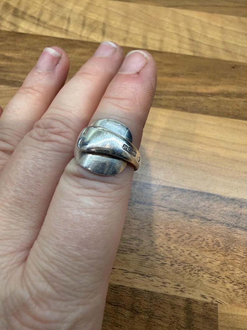 May include: A silver ring crafted from a spoon, featuring a curved design. The ring is worn on a finger, showcasing the repurposed silverware's unique form. The ring's surface reflects light, highlighting its metallic sheen and the repurposed craftsmanship.