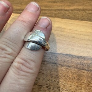 May include: A silver ring with a unique spoon-like design. The ring features a curved band that wraps around the finger, with a flattened, oval-shaped top. The ring is worn on a finger, showcasing its polished metal finish.
