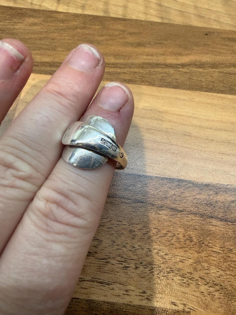 May include: A silver ring with a unique spoon-like design. The ring features a curved band that wraps around the finger, with a flattened, oval-shaped top. The ring is worn on a finger, showcasing its polished metal finish.