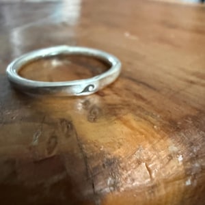 May include: A silver ring with a wave design. The ring is circular and has a smooth, polished surface. The wave design is a small, black graphic on the ring's surface. The ring is on a wooden surface.