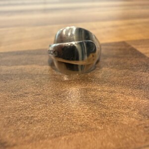 May include: A silver spoon ring with a curved design. The ring is made from a spoon and has a shiny, reflective surface. The ring is sitting on a wooden surface.