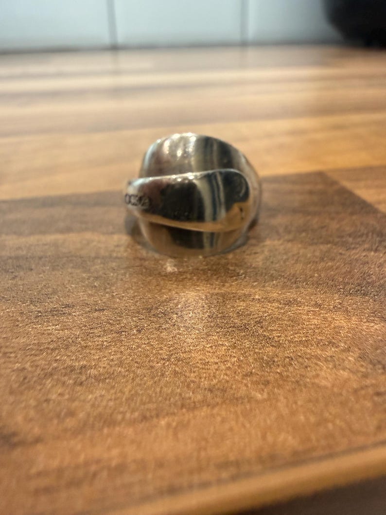 May include: A silver spoon ring with a curved design. The ring is made from a spoon and has a shiny, reflective surface. The ring is sitting on a wooden surface.