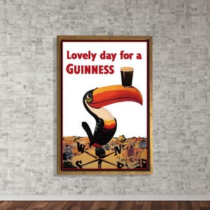 K&ouml;nnte beinhalten: Gerahmtes Vintage-Guinness-Poster mit einem Tukan, der ein Pint Guinness auf dem Schnabel balanciert. Der Text "Lovely day for a Guinness" ist in Rot und Wei&szlig;. Der Hintergrund zeigt eine Stadt und einen Kompass.