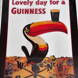 K&ouml;nnte beinhalten: Gerahmter Druck mit einem Tukan, der ein Guinness-Bier h&auml;lt. Der Tukan hat einen roten und orangen Schnabel, schwarz-wei&szlig;e Federn und den Text "Lovely day for a GUINNESS". Der Hintergrund zeigt eine Stadtlandschaft mit einem Kompass.