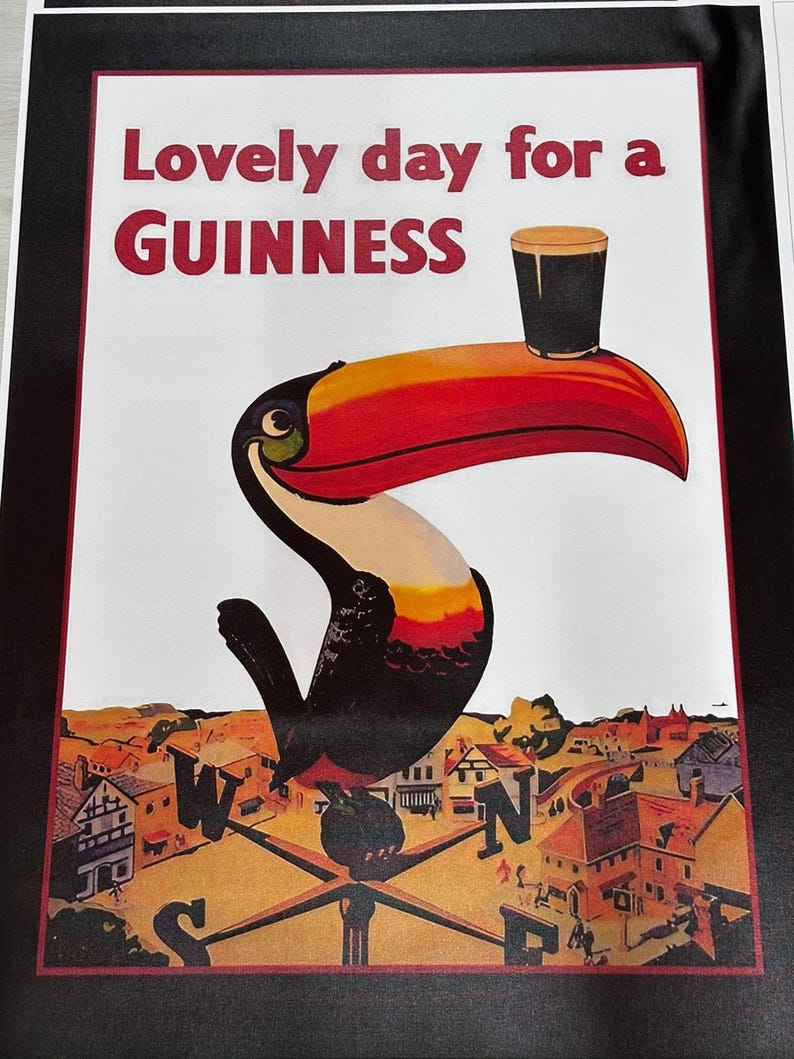 K&ouml;nnte beinhalten: Gerahmter Druck mit einem Tukan, der ein Guinness-Bier h&auml;lt. Der Tukan hat einen roten und orangen Schnabel, schwarz-wei&szlig;e Federn und den Text "Lovely day for a GUINNESS". Der Hintergrund zeigt eine Stadtlandschaft mit einem Kompass.