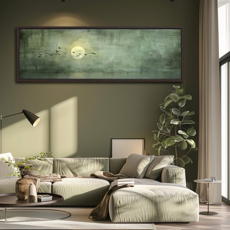 Puede incluir: Una gran obra de arte rectangular enmarcada representa un paisaje sereno. Una luna llena brilla intensamente, con p&aacute;jaros en vuelo sobre un fondo verde y gris apagado. La obra de arte cuelga sobre un sof&aacute; seccional verde claro en una sala de estar moderna.