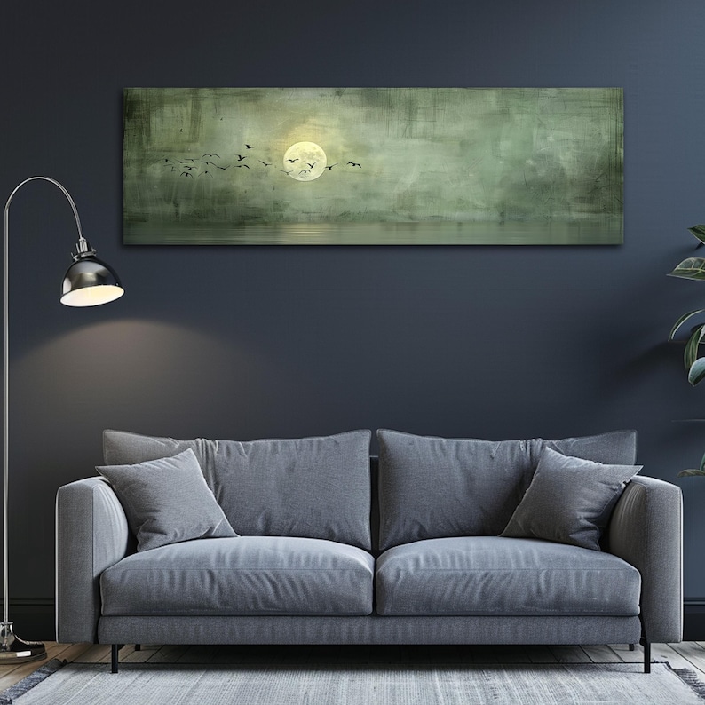 Puede incluir: Una obra de arte rectangular en lienzo representa una luna llena y p&aacute;jaros volando sobre un fondo verde y gris apagado. La obra cuelga sobre un sof&aacute; gris con cojines a juego. A la izquierda hay una l&aacute;mpara de pie plateada con una pantalla negra.