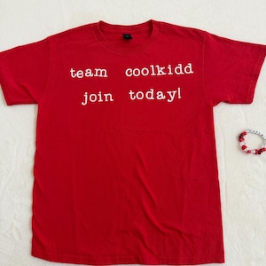 May include: A bright red t-shirt with the words "team coolkidd join today!" printed in white. A beaded bracelet with red and white beads is visible in the corner. The shirt is made of a soft material.