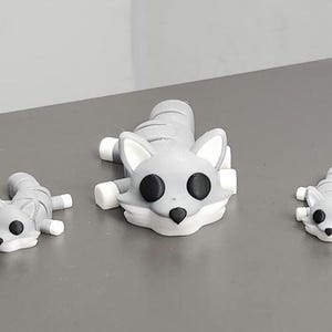 May include: Three gray and white fox figurines in various sizes. The foxes have black eyes and white accents on their ears, paws, and faces. The largest fox is lying down, while the smaller ones are in a similar pose.