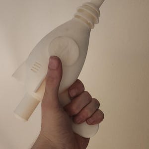 May include: A cream-colored toy ray gun, held in a hand. The ray gun has a bulbous body, a long barrel, and a handle. It features a circular detail and four parallel lines on the side.