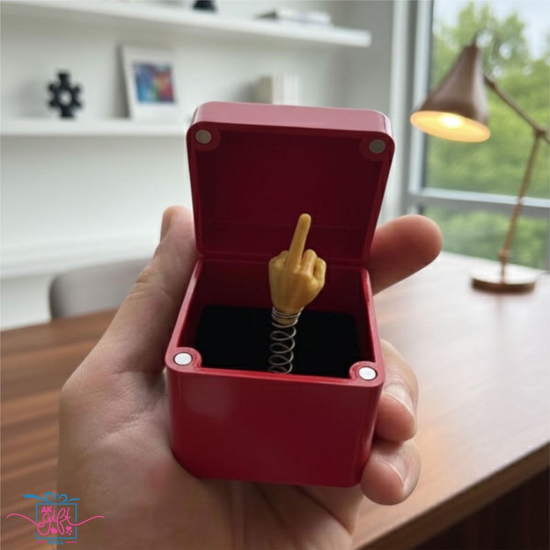 Op de afbeelding: Een rode vierkante doos met een scharnierend deksel, open om een gele hand te onthullen die de middelvinger opsteekt, gemonteerd op een zilveren veer. De doos wordt in een hand gehouden.