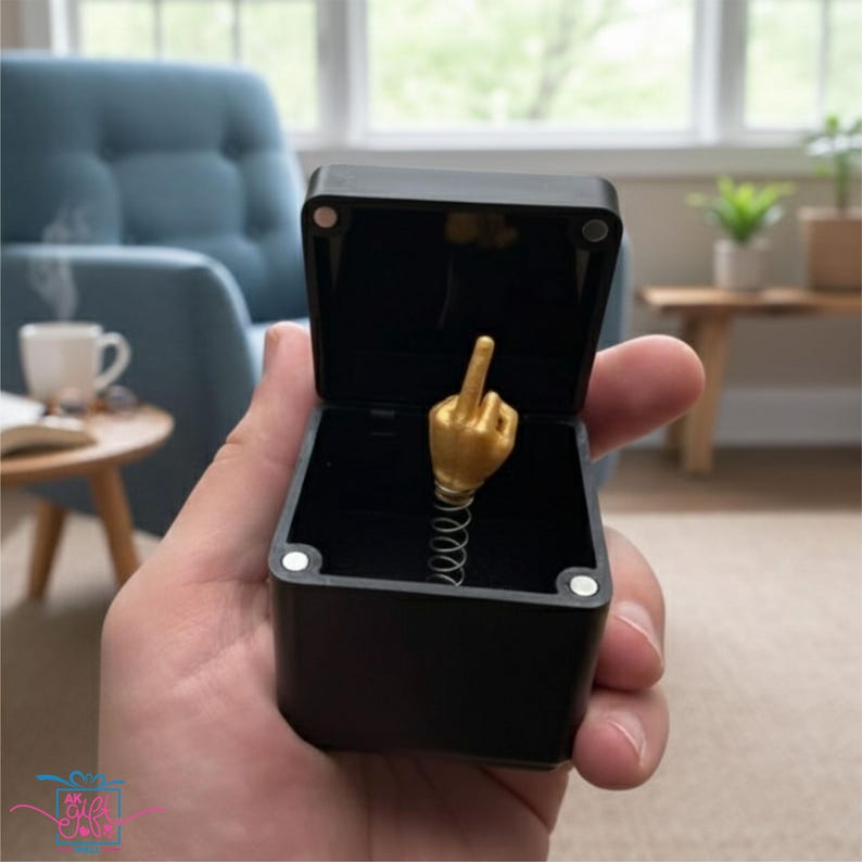 Op de afbeelding: Een open zwarte doos met een goudkleurige hand die de middelvinger opsteekt, gemonteerd op een veer. De achtergrond is wazig, met een glimp van een kamer.