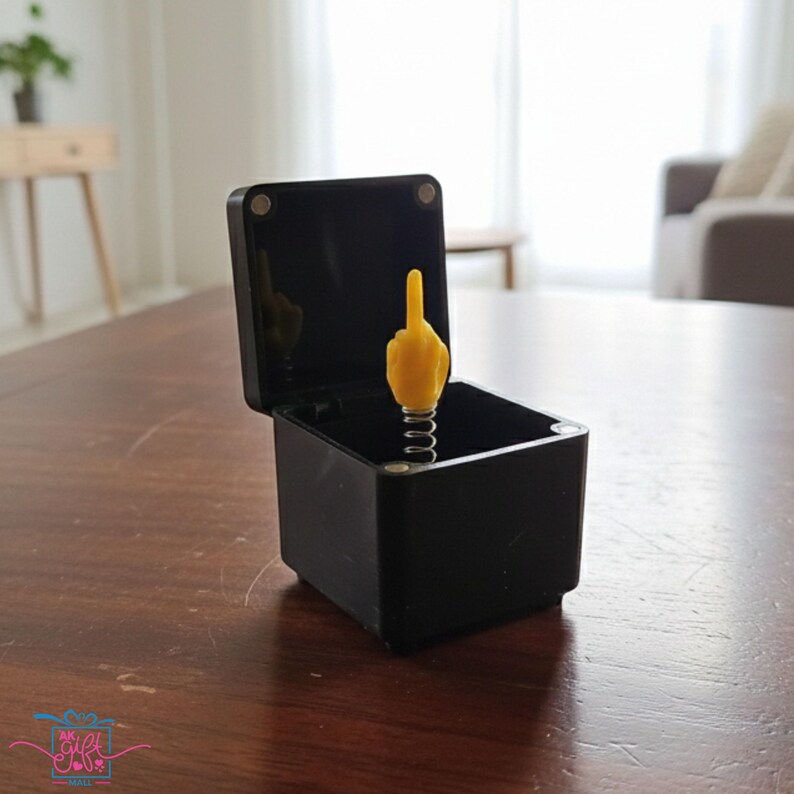 Op de afbeelding: Een zwarte, vierkante, open doos met een gele hand die de middelvinger opsteekt en uit een zilveren veer springt. De doos staat op een bruin houten oppervlak.