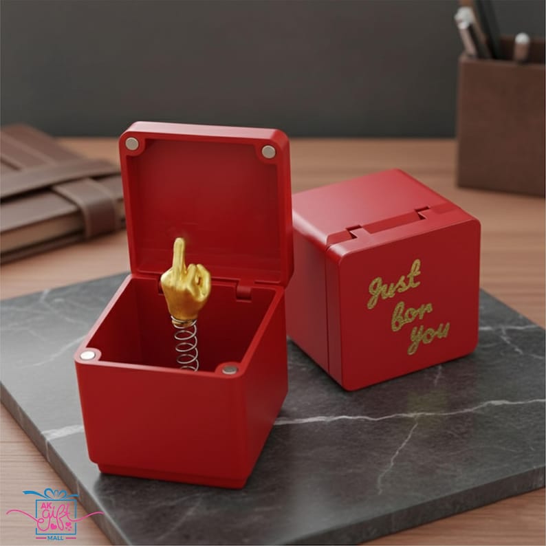 Op de afbeelding: Twee rode dozen, &eacute;&eacute;n open met een gouden hand die de middelvinger op een veer toont, de andere gesloten met de tekst "Just for you". De dozen staan op een marmerachtig oppervlak. De open doos heeft een vierkante vorm.