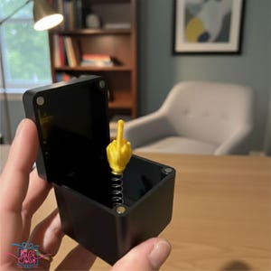 Op de afbeelding: Een zwarte doos met een gele middelvinger die uit een veer springt. De doos is open en onthult het novelty-item. De achtergrond is wazig en toont een kamer met een bureau, stoel en boekenkast.